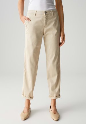 Beige cotton trousers with a relaxed fit, featuring back pockets, rolled cuffs, and a button closure, paired with beige slip-on shoes.