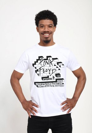Homme souriant portant un t-shirt blanc Pink Floyd avec un motif de mur en briques et des titres de chansons, debout les mains sur les hanches sur fond blanc.