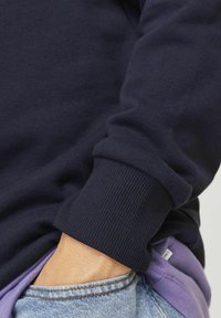 Sudadera azul marino con puños acanalados, sobre una camiseta morada. Piel visible en la muñeca, manos en un bolsillo de jeans.