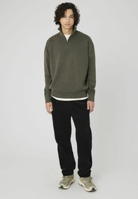 Pull à col zippé vert olive en tissu texturé, associé à un pantalon en velours côtelé noir et des baskets beiges, avec un t-shirt blanc visible en dessous.