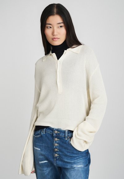 White ribbed knit pullover with a collar and button placket. Worn over a black turtleneck, paired with medium blue distressed jeans featuring button accents.