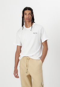 Hvit bomulls t-skjorte med rund hals, korte ermer, og liten svart logo på venstre bryst. Beige sweatpants med snøring og sidelommer.