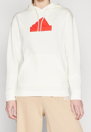 Osoba ubrana w biały kaptur z czerwonym logo Adidas oraz beżowe spodnie z małym białym logo Adidas.