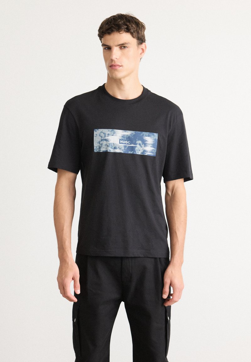 Ung mand iført en sort T-shirt med et blåt og hvidt rektangulært grafisk design og sorte bukser, der står foran en ensfarvet hvid baggrund.