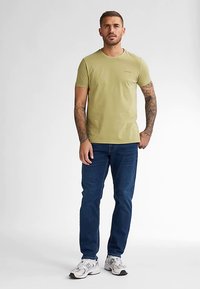 Oliivinvihreä puuvillainen t-paita, jossa on lyhyet hihat, pyöreä pääntie ja hillitty logodetalji. Yhdistetty sinisiin farkkuihin ja valkoisiin lenkkareihin.