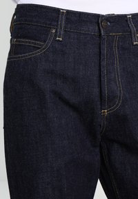 Dark blue denim jeans featuring yellow stitching, standard five-pocket design, and button closure at the waist. Smooth texture, tailored fit.