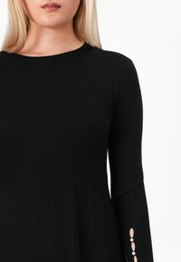 Femme portant un top noir en maille côtelée à manches longues avec des boutons en perles sur le poignet, montré du cou à la hanche sur fond blanc.