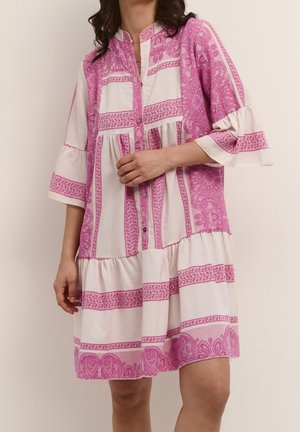 Kobieta ubrana w białą sukienkę do kolan z różowym wzorem paisley i pasków, z rękawami długości do łokcia oraz zapinana z przodu na guziki.