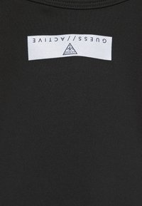 Tissu noir avec une étiquette rectangulaire blanche près de l'encolure affichant le texte inversé "GUESS // ACTIVE" et un petit logo triangulaire.