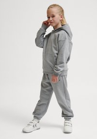 Grau Hoodie- und Jogginghosen-Set aus weichem Material, mit einer vorderen Tasche und Logodetails. Getragen mit weißen Sneakern.