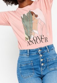 T-shirt rosa chiaro con una grafica che raffigura una donna, foglie di palma e il testo "LOVE AMOUR"; abbinato a jeans in denim blu a vita alta.