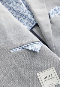 Blazer gris clair slim fit en tissu texturé ; doublure à motif bleu et pochette assortie ; étiquette de marque visible.