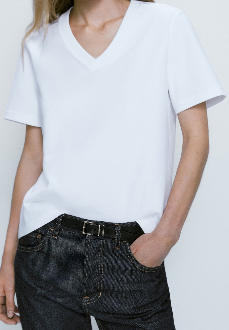 Jeune personne portant un t-shirt blanc uni à col en V et un jean foncé avec une ceinture noire, une main dans la poche sur un fond uni.