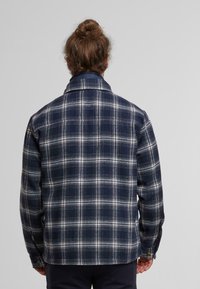 Camicia di flanella a scacchi blu navy e bianca con colletto e polsini a bottoni, caratterizzata da una texture morbida e un motivo a quadretti su tutta la superficie.