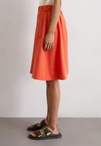 A mid-length, orange skirt with an elastic waistband and side pocket, paired with black sandals featuring gold buckles. Smooth texture.