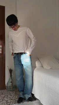 Mlad moški z očali, belim dolgim rokavom in modrimi kavbojkami stoji v notranjosti blizu postelje z belo posteljnino in stekleno vazo na tleh.