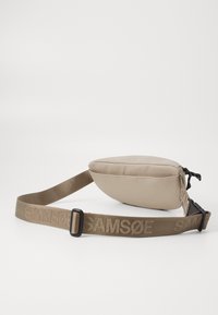 Beige színű nylon táska sima felülettel, lapos formával és állítható, hímzett márkanevével ellátott textil pánttal. Fekete cipzárhúzóval rendelkezik.