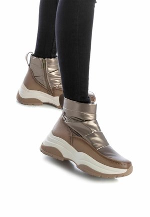 Botines de tobillo de bronce metálico con un acabado texturizado, que cuentan con una cremallera en el lateral, suelas gruesas blancas y un puño acolchado alrededor del tobillo.