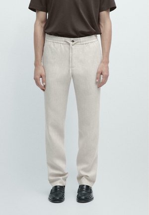 Mann trägt beige Kordelhose, braunes kurzärmeliges Hemd und schwarze Slipper, steht vor einfachem hellem Hintergrund.