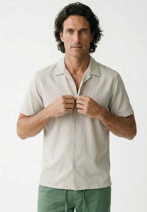 Hombre con cabello oscuro y ondulado que lleva una camisa beige claro de manga corta con botones y pantalones verdes, abotonándose la camisa.