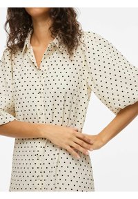 Femme aux cheveux bouclés portant une robe à manches courtes crème à boutons avec des pois noirs, mains croisées à la taille.