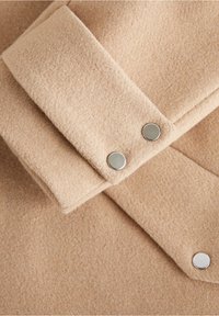 Tissu en laine beige avec une texture douce, comportant un col replié et deux boutons-pression en métal pour la fermeture.