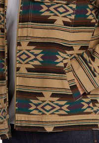Beige zip-up jacket with tribal patterns in brown, teal, and purple. Features a pocket on the left side and striped accents throughout.