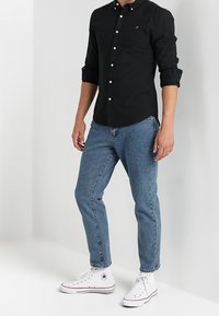 Hombre vistiendo una camisa negra de botones con las mangas arremangadas, vaqueros azules y zapatillas altas blancas, de pie contra una pared blanca.