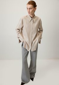 Bež košulja na pruge s dugmadi i kragnom, s širokim manžetama i srebrnim detaljima. Uparena s sivim širokim hlačama i crnim cipelama s oštrim vrhom.