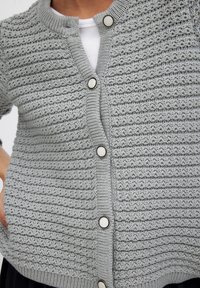 Šedý pletený cardigan s horizontálnym textúrovaným vzorom, okrúhlym výstrihom, predným zapínaním na gombíky a kontrastnými bielymi gombíkmi.