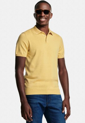 Lachende man met bruine zonnebril, geel poloshirt met korte mouwen en blauwe jeans, staand tegen een effen witte achtergrond.