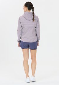 Kvinde, der står med ryggen til, iført en lys lilla hættejakke, mørkeblå shorts, hvide sneakers og en flettet hestehale.
