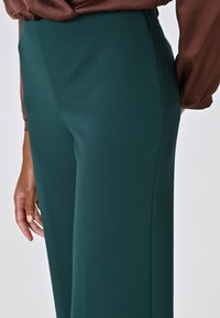 Pantalons taille haute teal en tissu lisse et extensible, présentant une coupe ajustée et des détails discrets sur les coutures latérales.