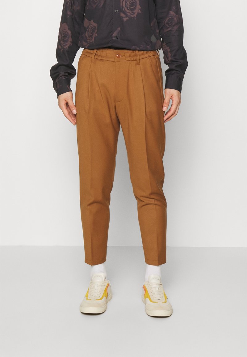 Homme portant un pantalon marron fuselé, des chaussettes blanches, des baskets avec des accents jaunes et une chemise noire à motifs rose foncé, debout sur un sol gris.