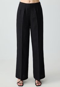 Pantaloni larghi in lino nero con pieghe frontali, vita alta e tasche laterali. Texture morbida, design lungo fino al pavimento e look minimalista.