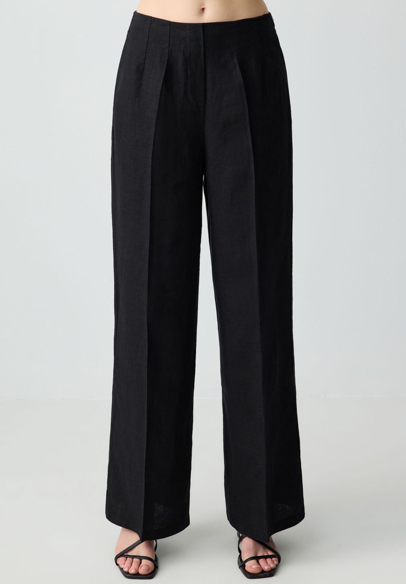 Pantaloni larghi in lino nero con pieghe frontali, vita alta e tasche laterali. Texture morbida, design lungo fino al pavimento e look minimalista.