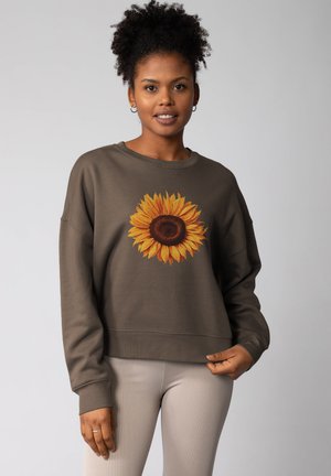 Brauner Sweatshirt mit einem großen Sonnenblumenmotiv in Gelb und Braun, mit lockerer Passform und gerippten Bündchen. Kombiniert mit hellen gerippten Leggings.
