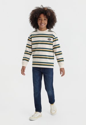 Uśmiechnięte dziecko z kręconymi włosami, ubrane w biały, zielono-żółty pasiasty sweter, niebieskie dżinsy i białe trampki, stojące na jednolitym tle.