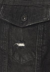 Gros plan sur un tissu en denim noir avec un rabat de poche boutonné et une petite déchirure rectangulaire usée sous la poche.