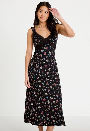 Kvinde i en sort blomsterprintet ærmeløs midikjole med blondenederdel, stående mod en ensfarvet hvid baggrund, mens hun rører ved sit hoved.