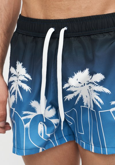 Maillot de bain bleu avec cordons blancs et imprimé de palmiers blancs porté par une personne montrant la taille et la partie supérieure des cuisses.