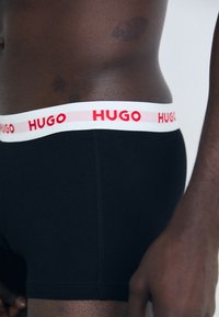 Schwarze Boxershorts mit einem weißen Bund, der mit rotem "HUGO"-Text versehen ist. Der Stoff erscheint glatt, mit einer engen Passform und ohne sichtbare Muster.