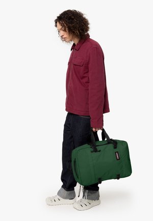 Green bag with black accents, rectangular shape, fabric material. Model wears a maroon shirt and dark denim with sandals.