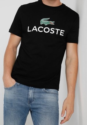 Černé bavlněné tričko s krátkými rukávy, na kterém je zelené logo krokodýla a bílý text "LACOSTE". Nosí se s světle modrými džíny.