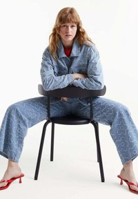 Chaqueta de denim azul claro y pantalones con bordado floral, combinados con sandalias de tacón rojas. Modelo sentado en una silla negra con los brazos cruzados.