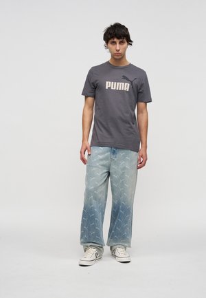 Młody mężczyzna stoi twarzą do przodu, ma na sobie szarą koszulkę PUMA, jasnoniebieskie szerokie dżinsy z wzorem oraz białe sneakersy w prostym studio.