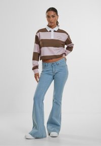 Crop top med brune og rosa striper, hvit krage, kombinert med lyseblå vide jeans og hvite joggesko. Enkelt logodesign på toppen.