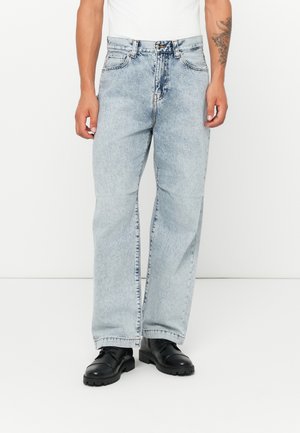 Hellblau gefärbte Jeans im Straight-Leg-Stil mit einem verwaschenen Finish, fünf Taschen und einem Knopfverschluss, kombiniert mit schwarzen Schnürschuhen.