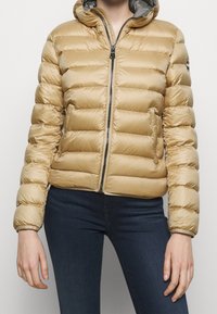 Crèmekleurige pufferjas met horizontale stiksels, voorzien van een ritssluiting aan de voorkant, een capuchon en een gladde, glanzende textuur. Draagt over donkere spijkerbroek.