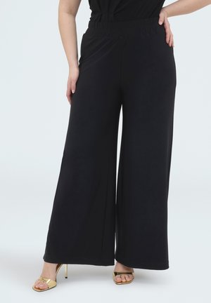 Kvinde iført sorte bukser med vide ben og gyldne højhælede sandaler, stående med den ene hånd på hoften mod en hvid baggrund.